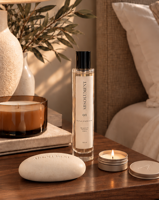Bouteille de parfum et mini bougie parfumée 01 d'Absolument Bergamote, thé vert et cèdre et galet à parfumer crème minéral déposés sur une table de chevet en bois près d'un lit en lin