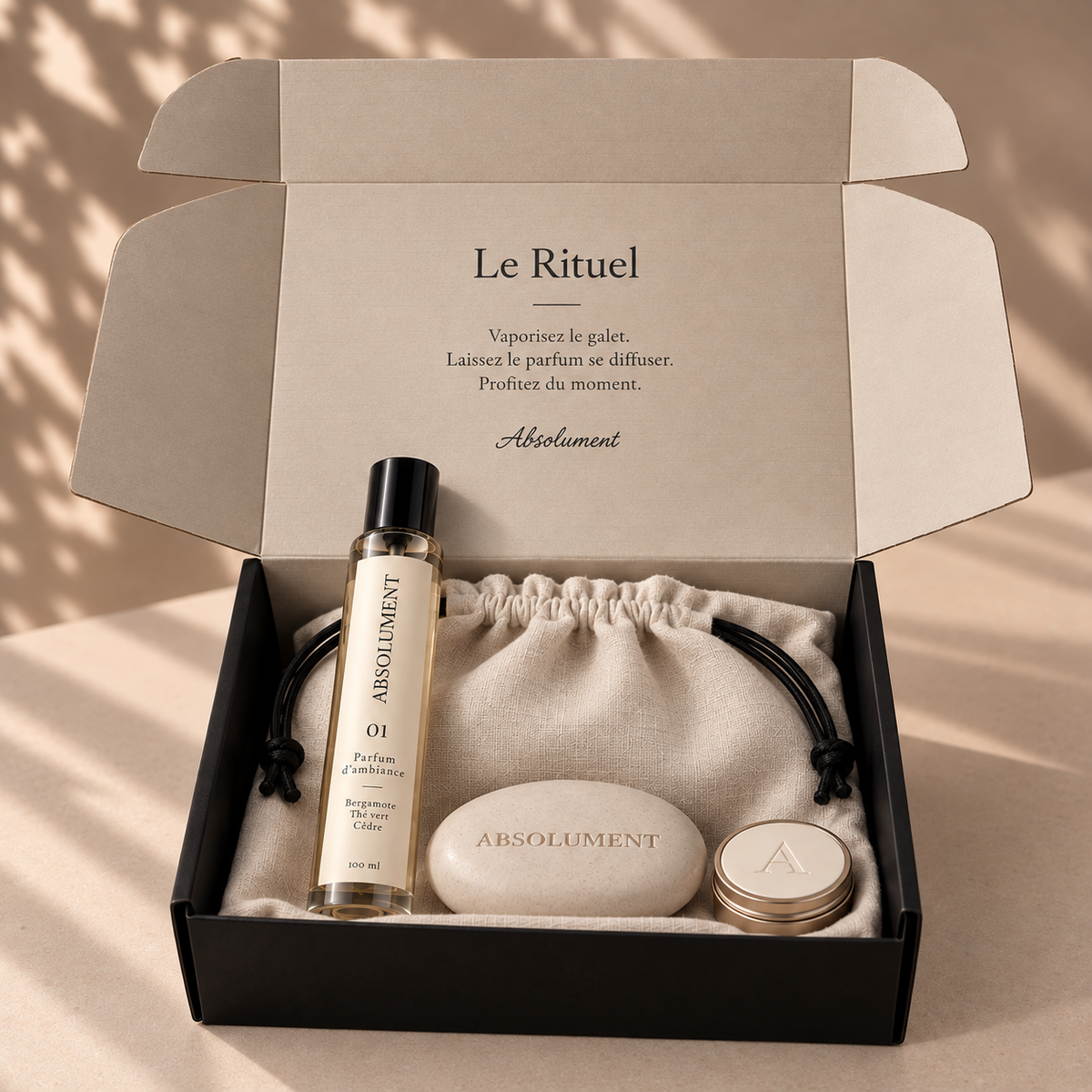 Coffret cadeau Le Rituel d’ABSOLUMENT, présentant le parfum d’ambiance 01 de 100 ml, un galet minéral crème, une mini bougie et une pochette en lin naturel dans une boîte noire au couvercle crème.